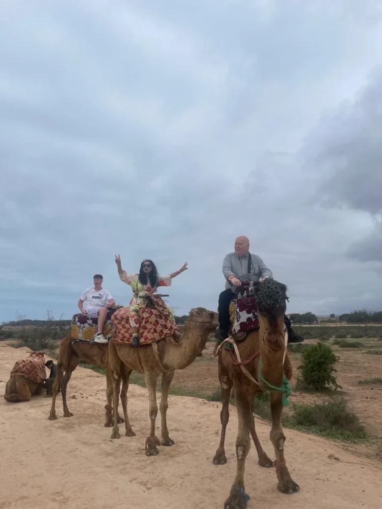 Three visitors on camels standing on sandy ground under an overcast sky