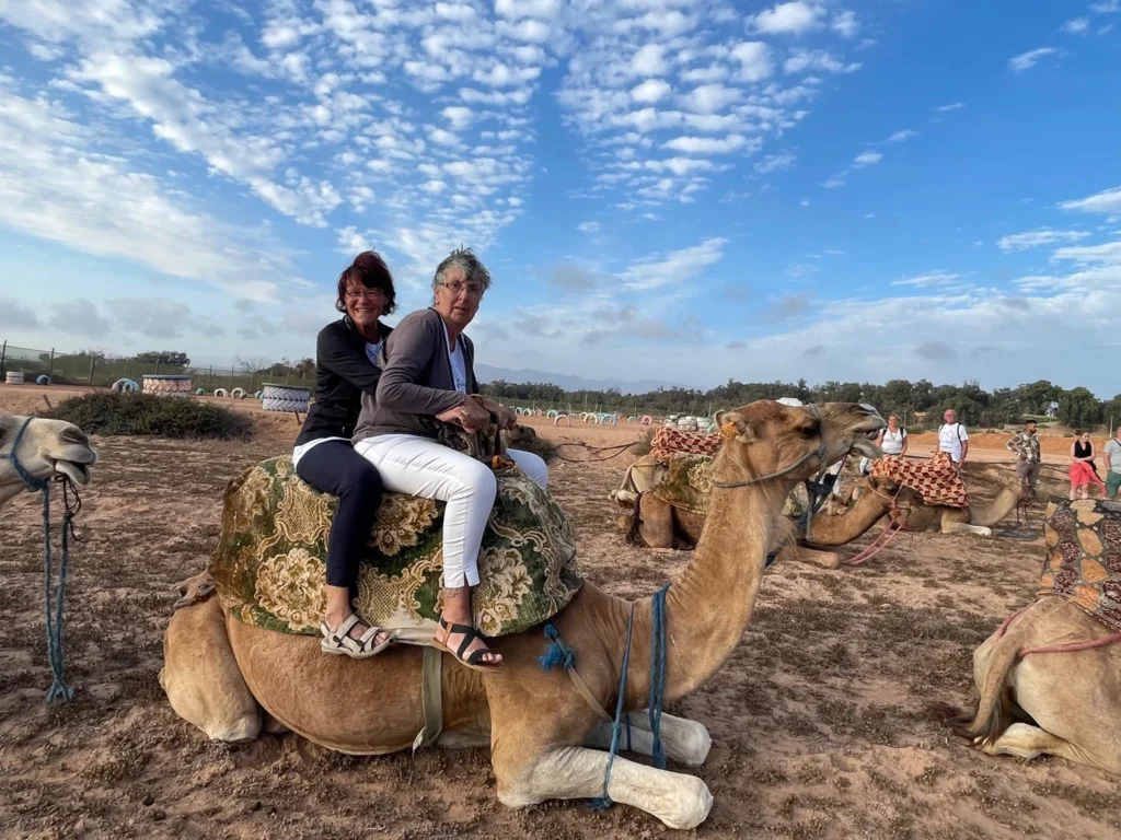 Two visitors seated together on a resting camel with other camels around