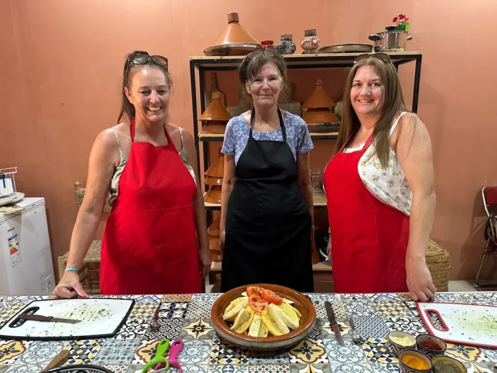 Three guests in aprons beside a finished tagine dish on a decorated tile table