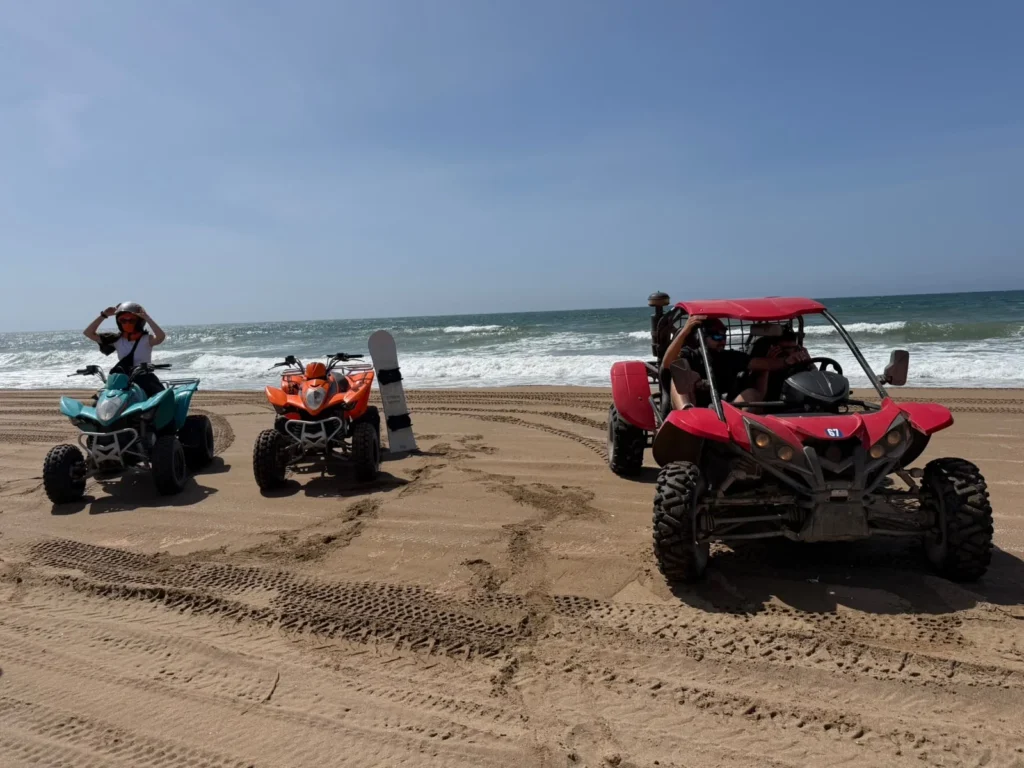 Quad bikes and a red buggy on the beach with a sandboard standing in the sand