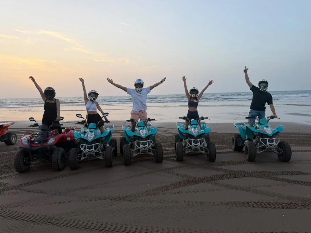 Five riders standing on quad bikes on a beach with their arms raised at sunset