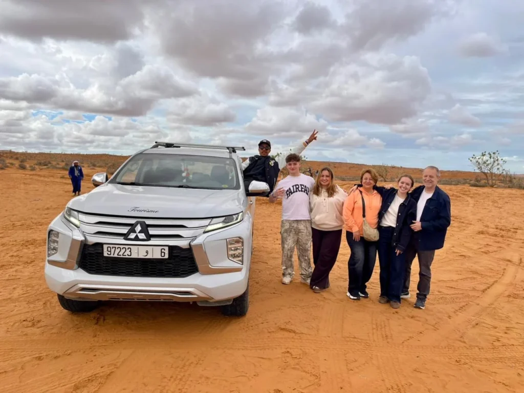 White 4x4 jeep parked on orange sand with a group of visitors standing beside it